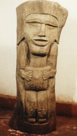 Figura de ndio esculpida em madeira escura no formato cilindrco, parte superior em incavo terminando em cocar. 
